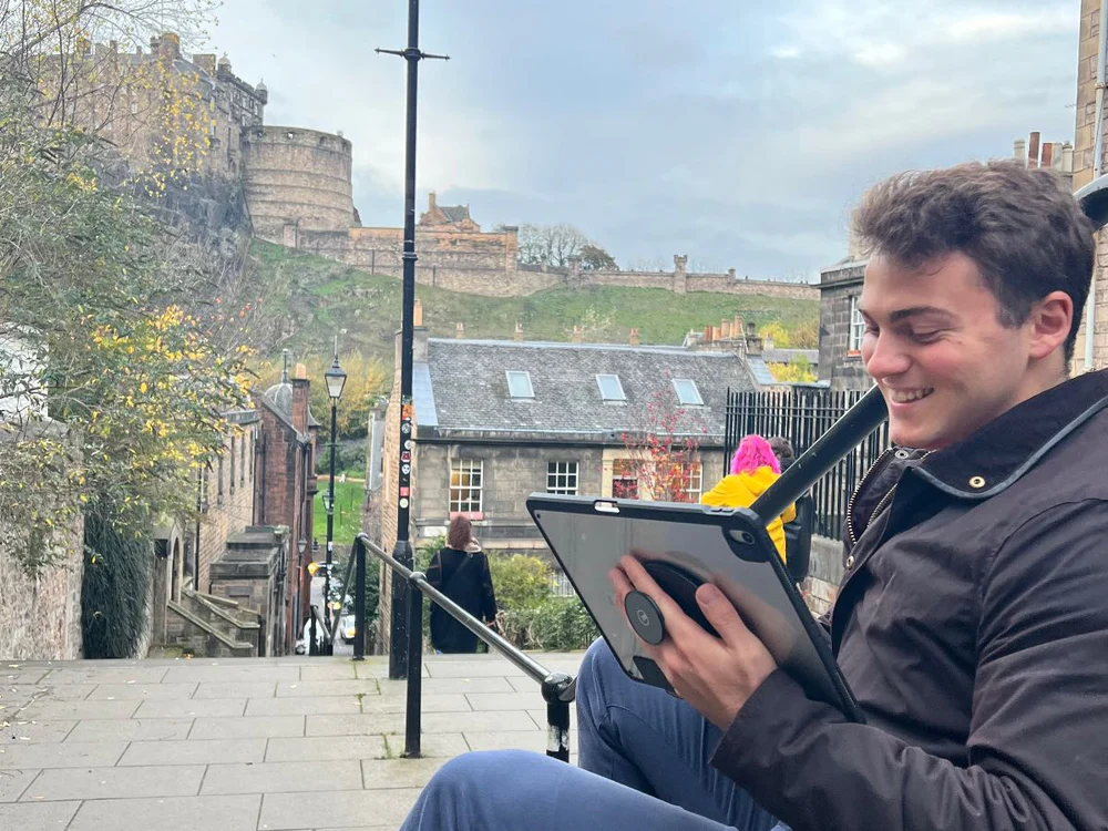 A digital nomad sits at The Vennel in Edinburgh with a view of Edinburgh in the background. He is holding an iPad with a G-Hold on the back of it, smiling as he works.